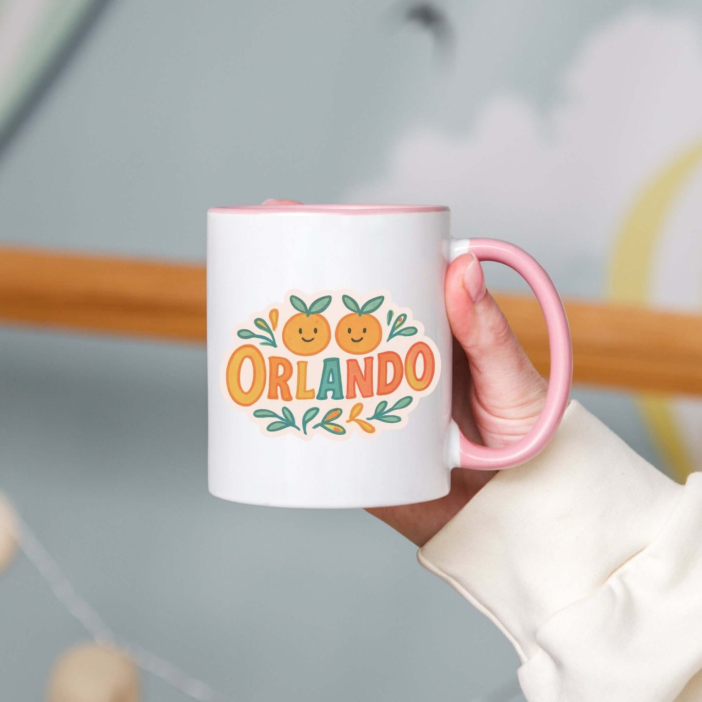 A person holding a white ceramic mug with 'Orlando' written in orange lettering, accompanied by a graphic of orange and green leaves. The mug has a pink interior and handle.