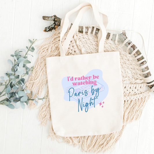 A white canvas tote bag with the text 'I'd Rather be watching Paris by Night' printed in blue and pink. The bag is displayed on a white surface with a decorative item and some greenery in the background.