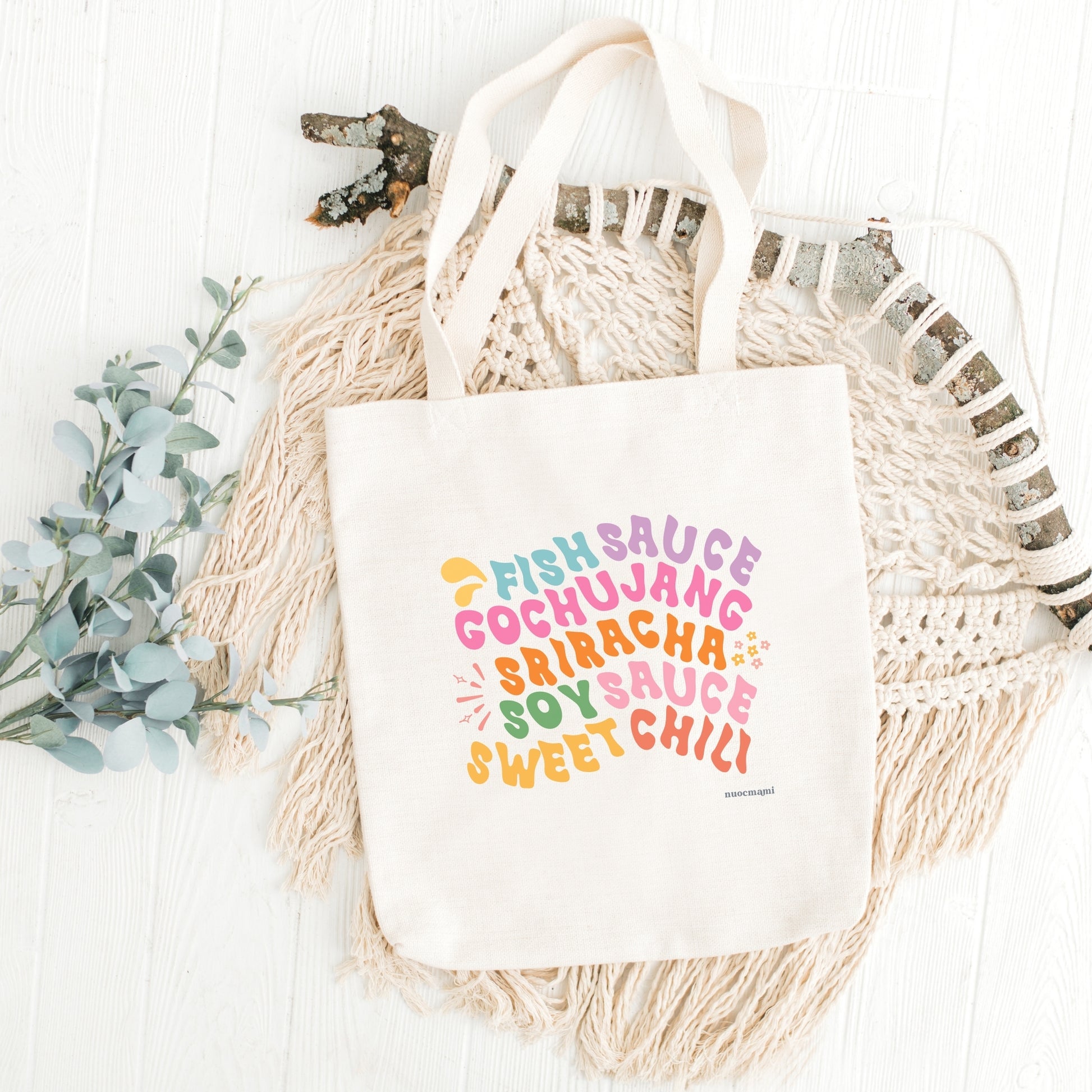 A white canvas tote bag with multicolored text printed on it, listing 'Fish Sauce', 'Gochujang', 'Sriracha', Soy Sauce', and 'Sweet Chili ', displayed on a surface with a decorative item to the left.