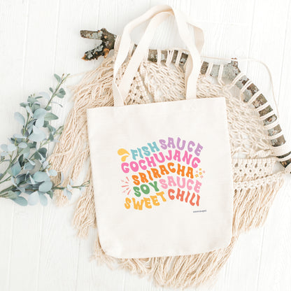 A white canvas tote bag with multicolored text printed on it, listing 'Fish Sauce', 'Gochujang', 'Sriracha', Soy Sauce', and 'Sweet Chili ', displayed on a surface with a decorative item to the left.