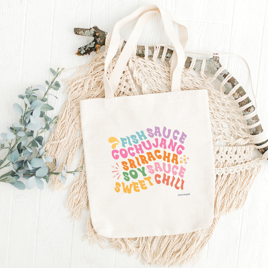 A white canvas tote bag with multicolored text printed on it, listing 'Fish Sauce', 'Gochujang', 'Sriracha', Soy Sauce', and 'Sweet Chili ', displayed on a surface with a decorative item to the left.