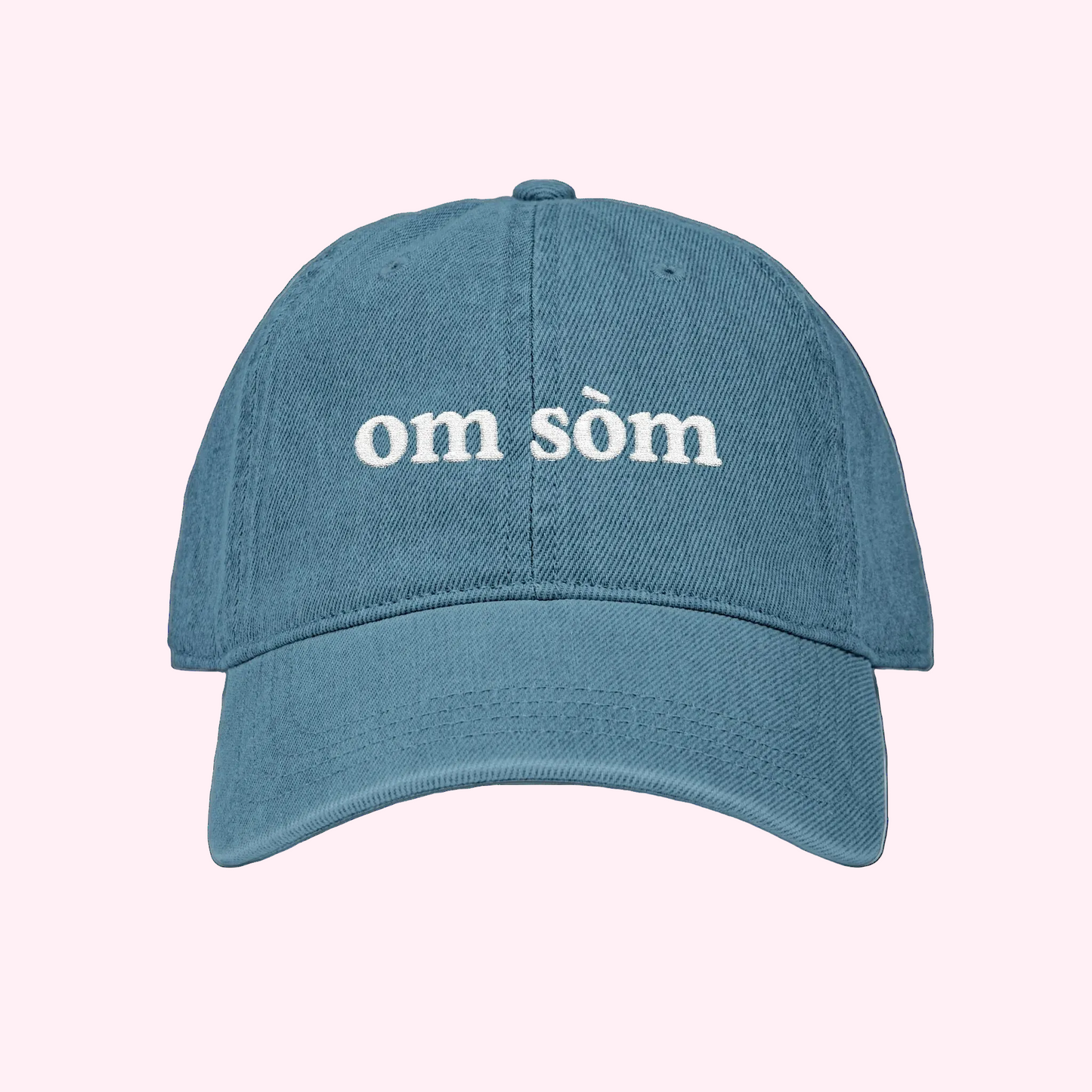 A blue denim-style baseball cap with 'om sòm' embroidered in white on the front, shown against a plain white background.
