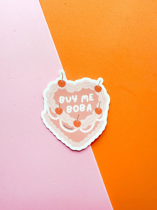 A matte premium vinyl sticker with the phrase 'BUY ME BOBA' written on a heart shaped cherry on top cake