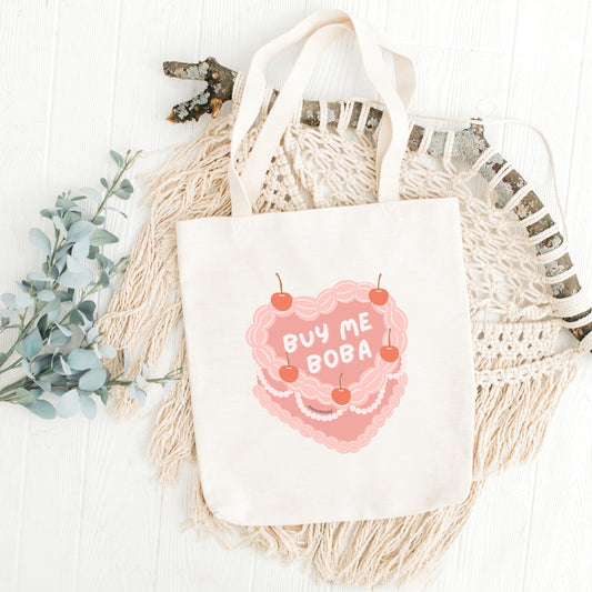A white canvas tote bag with 'BUY ME BOBA' text and a heart symbol printed on the front, displayed on a white background with decorative elements.