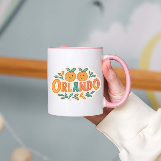 A person holding a white ceramic mug with 'Orlando' written in orange lettering, accompanied by a graphic of orange and green leaves. The mug has a pink interior and handle.
